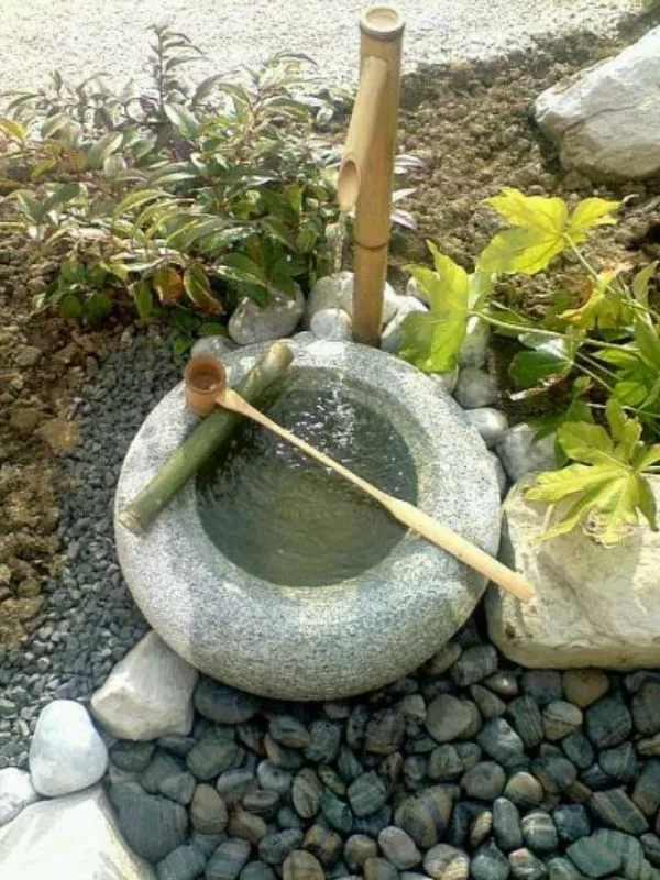 Fontaine zen en bambou et bassin en pierre tsukubai sur un lit de galets gris.